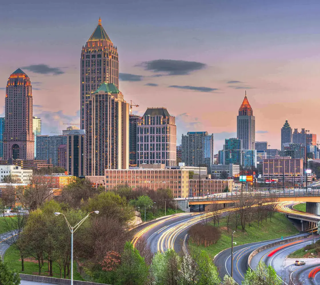 Atlanta_Skyline_Abendverkehr.jpg Skyline von Atlanta, USA, bei Sonnenuntergang mit beleuchteten Hochhäusern und fließendem Abendverkehr auf den geschwungenen Highways im Vordergrund.
