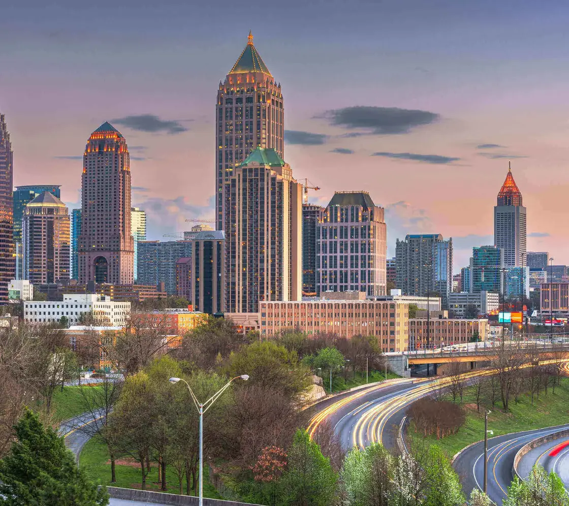 Atlanta_Skyline_Abendverkehr.jpg Skyline von Atlanta, USA, bei Sonnenuntergang mit beleuchteten Hochhäusern und fließendem Abendverkehr auf den geschwungenen Highways im Vordergrund.