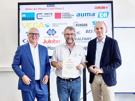 Sutter_WVIB-Robotiktag_2025_Übergabe_Stiftungsurkunde Drei Männer bei der Urkundenübergabe des WVIB Robotiktags 2025, im Hintergrund Sponsorenlogos auf einer Präsentationswand.