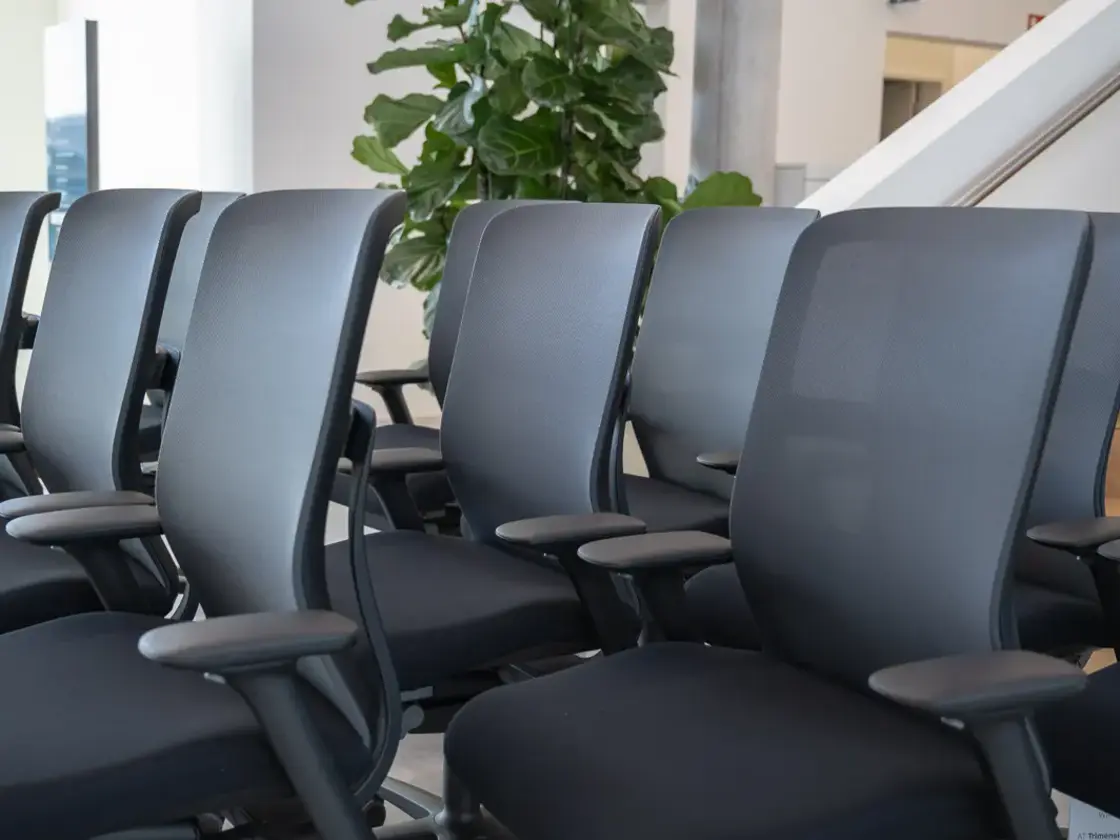 Reihe schwarzer Bürostühle mit Netzrücken und Armlehnen in einem Büro oder Konferenzraum, im Hintergrund große grüne Pflanze und Treppe