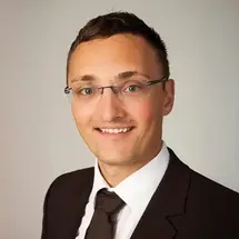 Heiser mit Brille, Hemd und Krawatte vor hellem, neutralem Hintergrund.