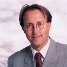 Dr. Brehmer mit Brille, grauem Anzug und roter Krawatte vor hellem Hintergrund.