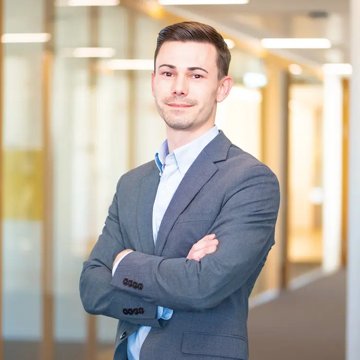 Ein junger Mann im grauen Anzug steht mit verschränkten Armen im modernen Büroflur von Sutter Medizintechnik und blickt selbstbewusst in die Kamera.