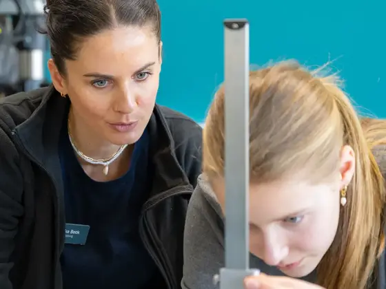 Junge Teilnehmerin des Girls’Day misst mit Unterstützung einer Sutter-Mitarbeiterin ein Werkstück in der Fertigung; beide konzentriert bei der Arbeit vor türkisfarbener Wand.