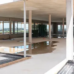 emmendingen-gebaeude-innenausbau-2021.jpg Blick in den Innenausbau des Sutter-Neubaus in Emmendingen mit großen Fensterflächen, Betonstützen und abgestellten Glasscheiben. Pfützen auf dem Boden spiegeln das Tageslicht.