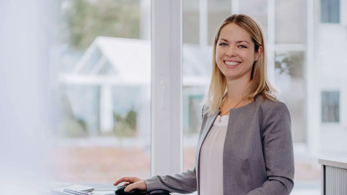 Sutter-Mitarbeiterin-Einkauf.jpg Eine Mitarbeiterin des Einkaufsteams steht lächelnd am höhenverstellbaren Schreibtisch und arbeitet am Computer.