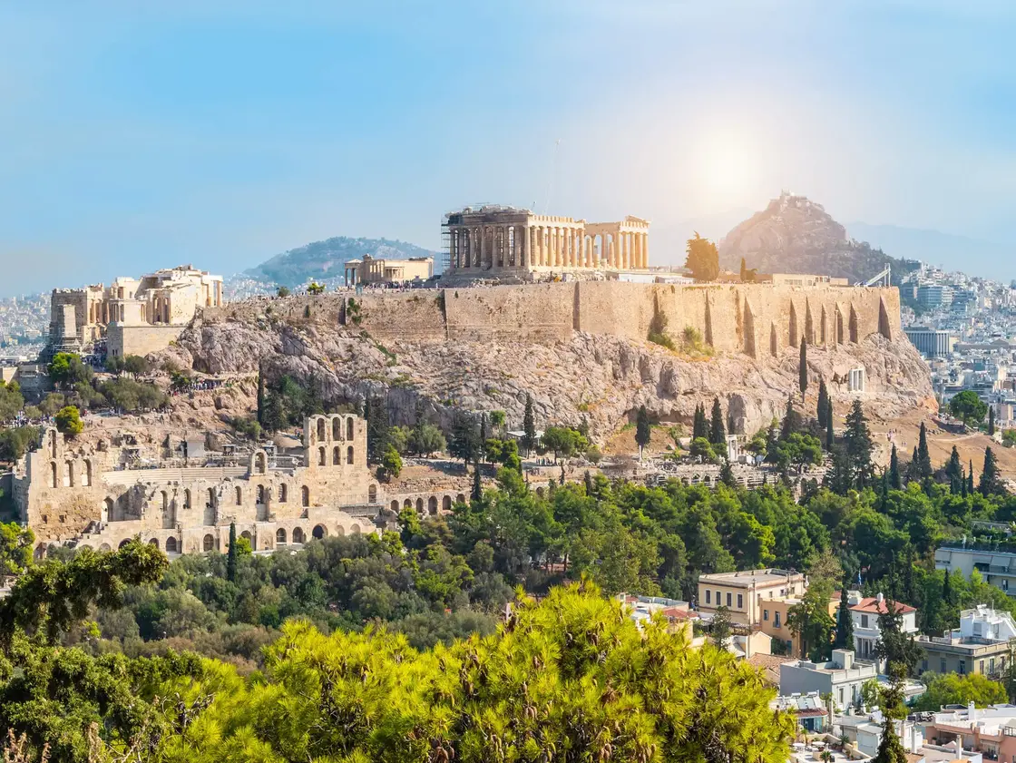 Athen_Akropolis_Parthenon_Header.jpg Blick auf die Akropolis in Athen mit dem Parthenon-Tempel und umliegenden antiken Bauwerken – im Hintergrund moderne Stadtlandschaft und grüne Hügel.