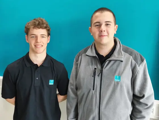 Zwei Personen mit Firmenkleidung und lächelnd vor türkisfarbenem Hintergrund